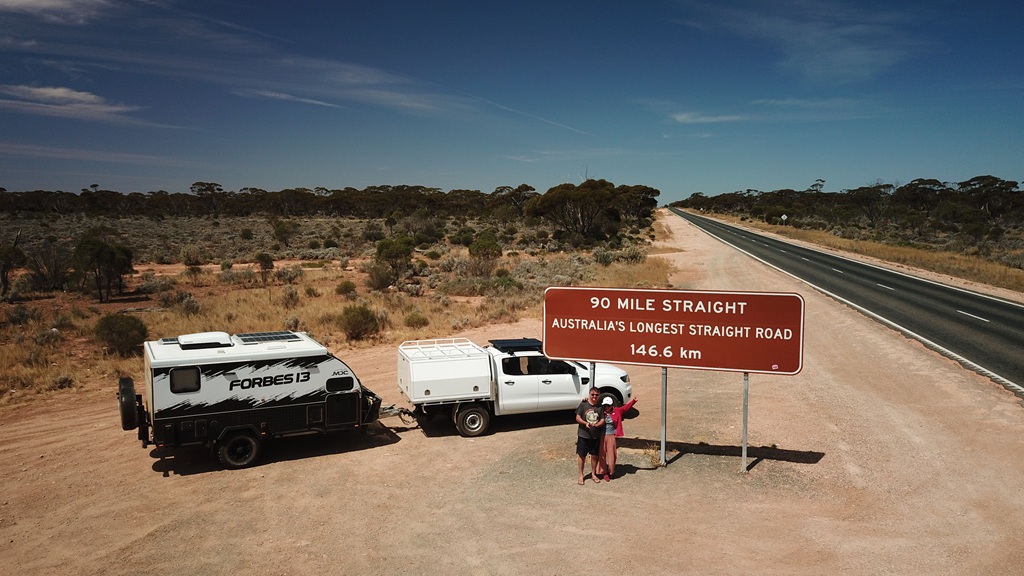 Rob and Muay standing by the 90 mile straight sign on the Nullarbor with tow vehicle and MDC hybrid caravan.