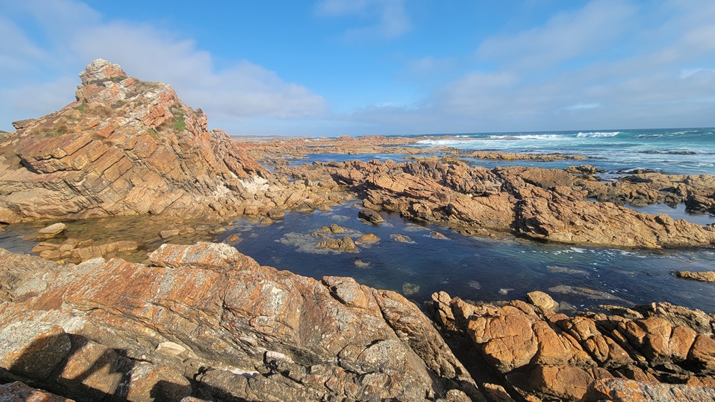 Jagged, orange-hued rock formations and clear tidal rock pools along the wild Arthur River coastline, with the Southern Ocean and a blue sky in the background.
