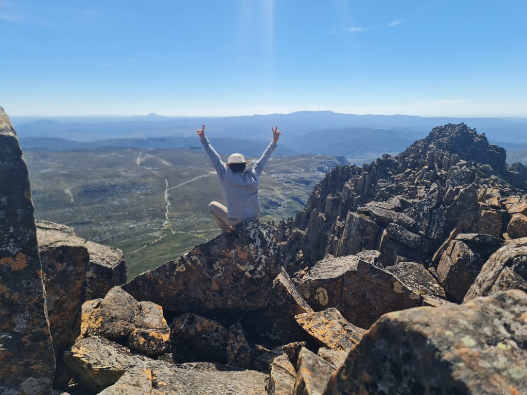 Hiker celebrating at the summit of Cradle Mountain with arms raised overlooking the Tasmanian Wilderness.