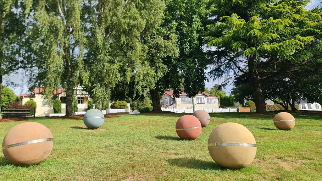 Large, colorful spherical stone sculptures with metallic bands situated on a grassy park lawn along the Deloraine Sculpture Trail, with heritage houses and mature trees in the background.