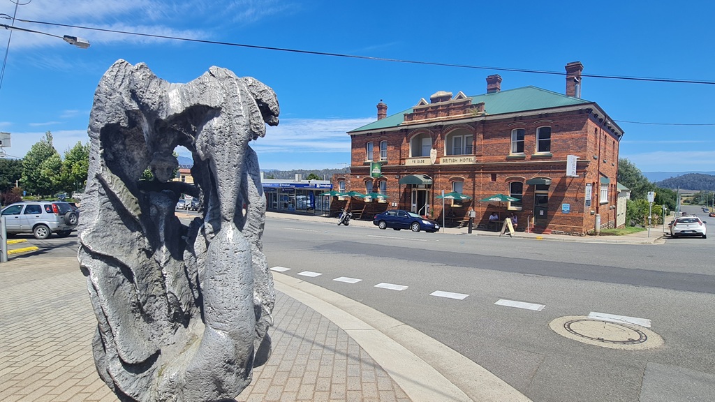 A large grey abstract street sculpture in the foreground with the historic red-brick Ye Olde British Hotel in the background. Located on the heritage streetscape of Deloraine, Tasmania, under a clear blue sky