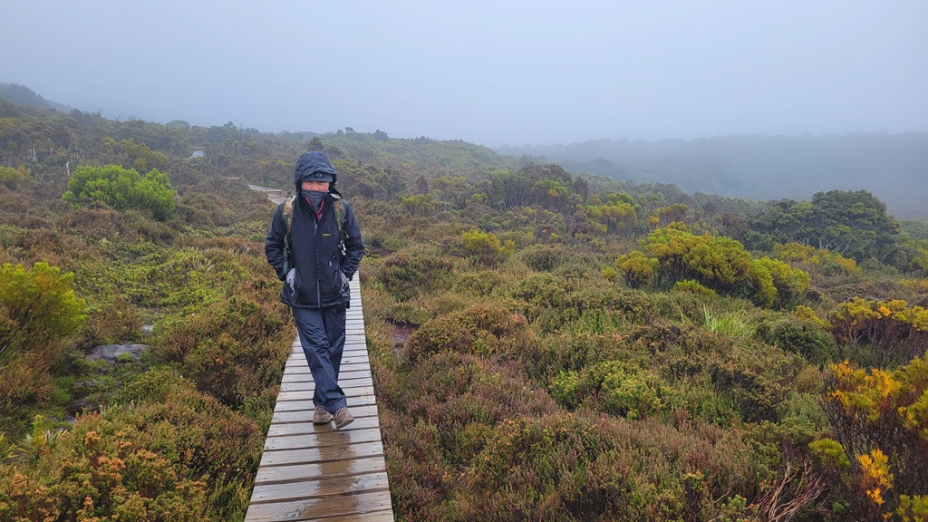 A woman hiker, Muay, heavily layered in cold-weather gear, walking through the misty and wet wilderness of Hartz Mountains National Park, Tasmania.