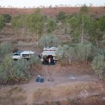 Aerial drone view of a remote off-grid campsite in the Australian outback featuring a 4WD and small caravan setup with solar panels and a Starlink dish deployed in a vast, empty landscape.
