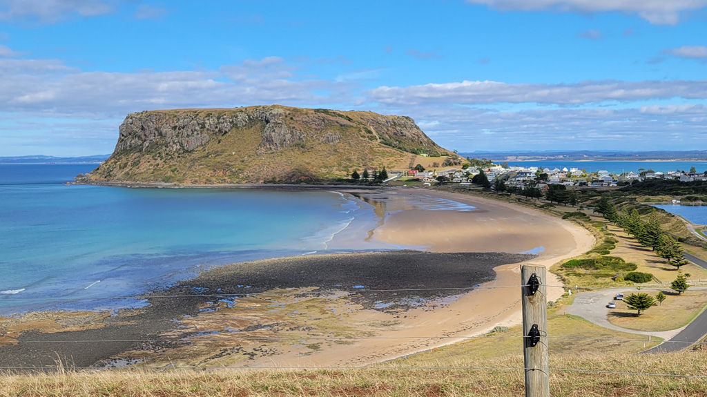 A wide-angle landscape view of The Nut, a massive flat-topped volcanic plug, rising above the turquoise waters of Bass Strait on a clear sunny day. The historic village of Stanley sits nestled at its base, viewed from the elevated green pastures near Highfield Historic Site. It is a fitting end of a Launceston or Devonport road trip.