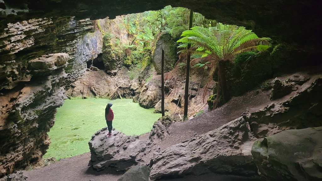 A view through the massive limestone Trowutta Arch in the Tarkine, looking out onto a vibrant green sinkhole pond with Muay standing on the rocks in the foreground to show the scale of the natural formation.