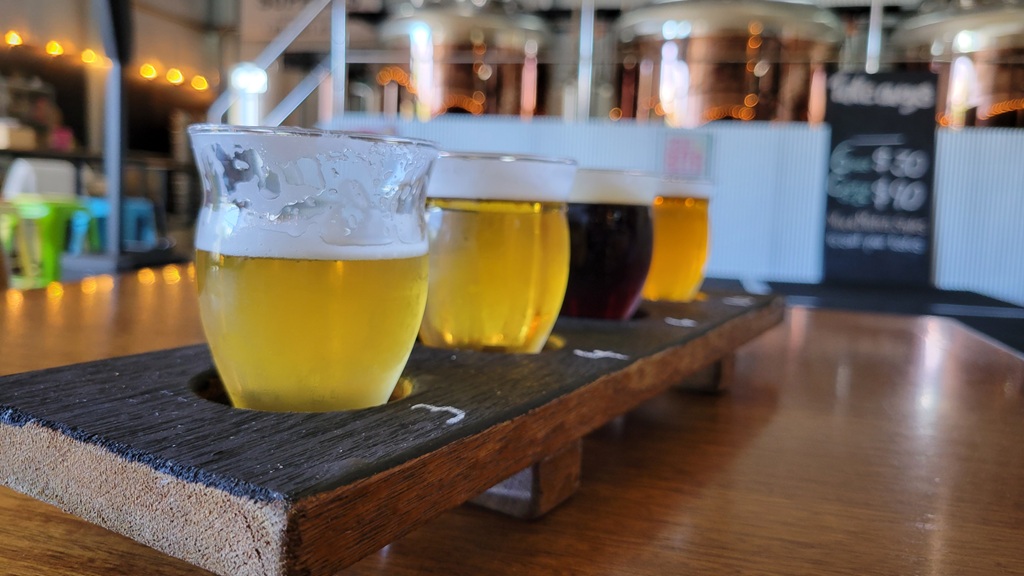 A close-up of a rustic wooden beer paddle with four different craft beer tasters on a table at a Barossa Valley brewery, with the industrial brewing tanks softly blurred in the background during a South Australia road trip.