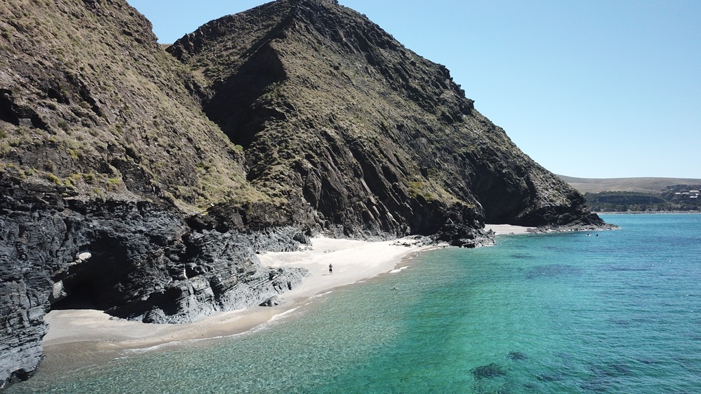 A drone photograph capturing the rugged coastal scenery at Rapid Bay on the Fleurieu Peninsula during a South Australia road trip. The image features a solitary figure on a secluded white sand beach, bordered by massive dark rocky cliffs, a coastal cave, and clear vibrant turquoise water.