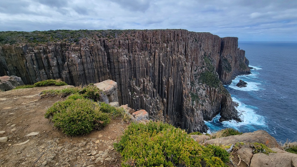 Wide-angle view of the vertical dolerite columns at Cape Raoul, Tasmania, showing the sheer cliffs dropping into the churning blue Southern Ocean under a cloudy sky.