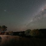 A wide-angle night photograph showing the Milky Way galaxy stretching across a clear, star-filled sky. In the foreground, silhouetted trees and tall grass frame a dark mountain range, with a small, glowing campsite and vehicle visible on the far left.