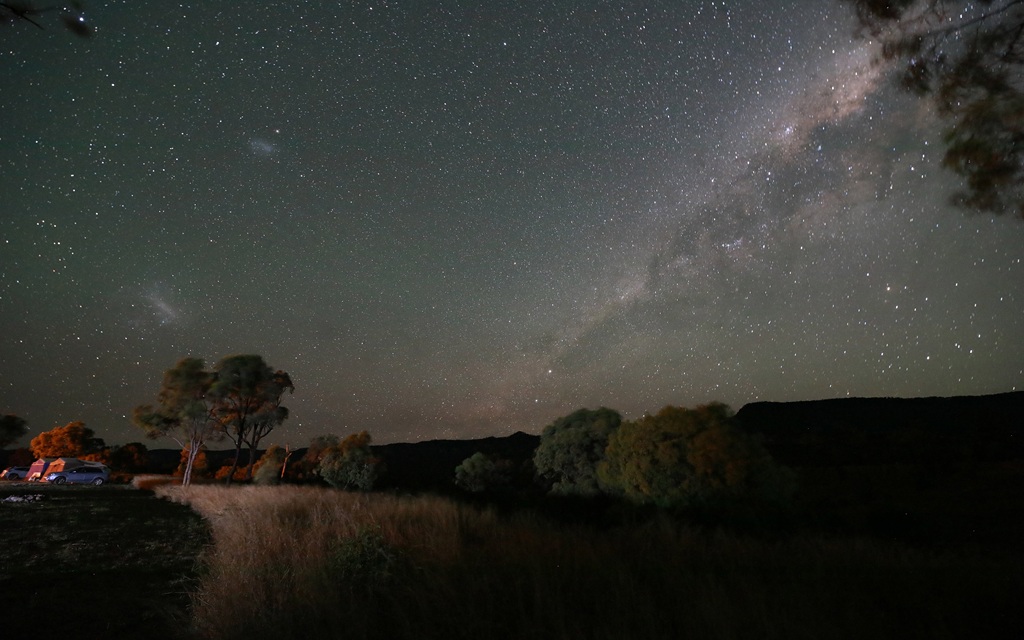 A wide-angle night photograph showing the Milky Way galaxy stretching across a clear, star-filled sky. In the foreground, silhouetted trees and tall grass frame a dark mountain range, with a small, glowing campsite and vehicle visible on the far left.