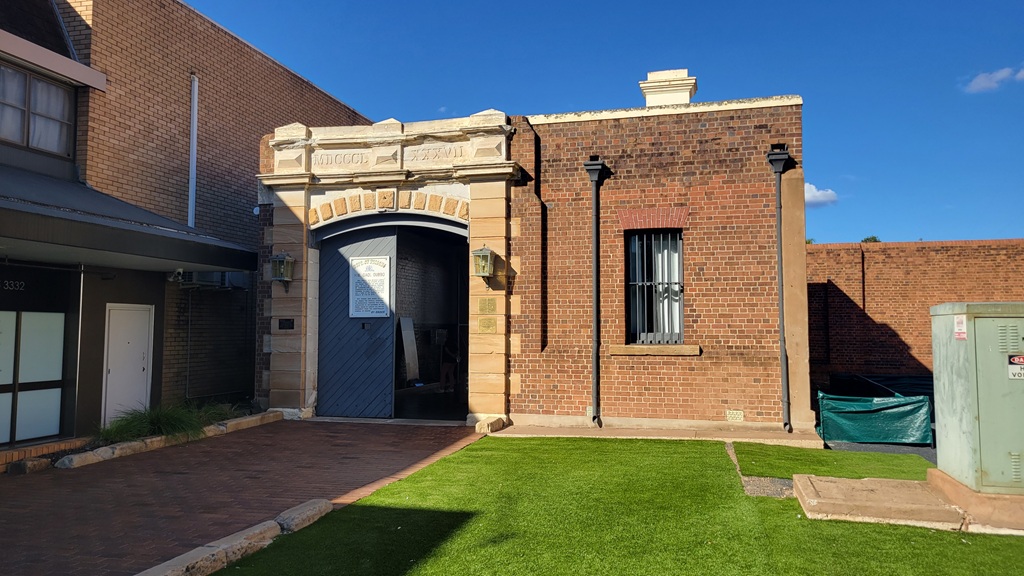 Historic entrance to Old Dubbo Gaol on the Great Inland Way, featuring 19th-century brickwork and large wooden gates under a clear blue sky.