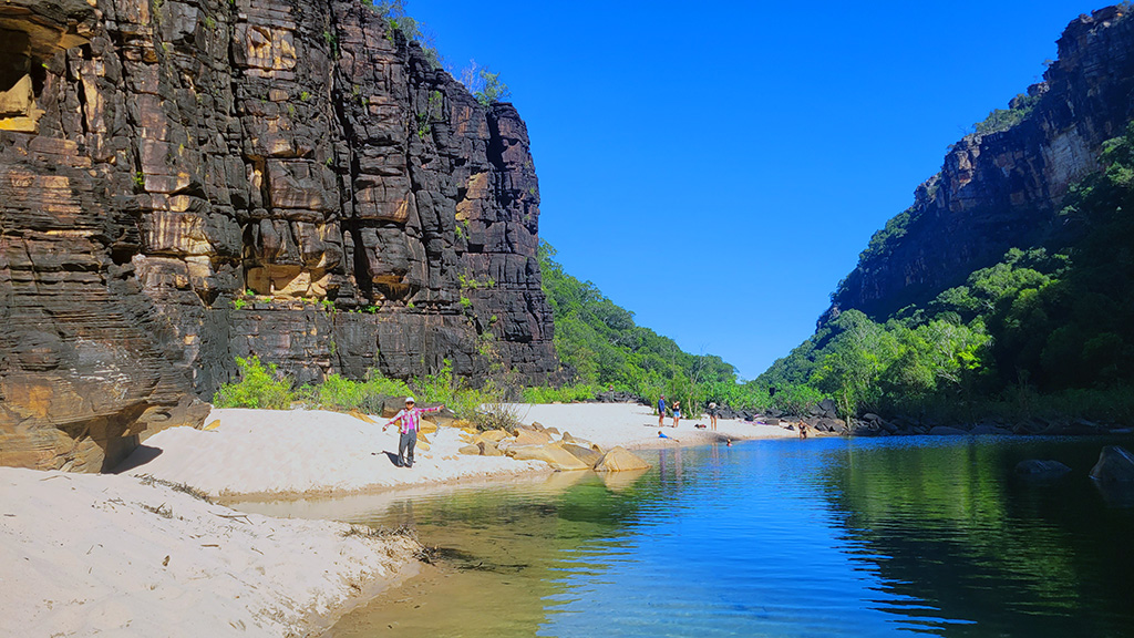 A woman standing on the white sand beach at the base of the Jim Jim Falls plunge pool, surrounded by towering dark rock cliffs and clear blue water.