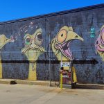 Great Inland Way QLD A large, quirky outdoor mural in Lightning Ridge featuring five stylized, expressive emu heads painted on a black brick wall at a local service station.