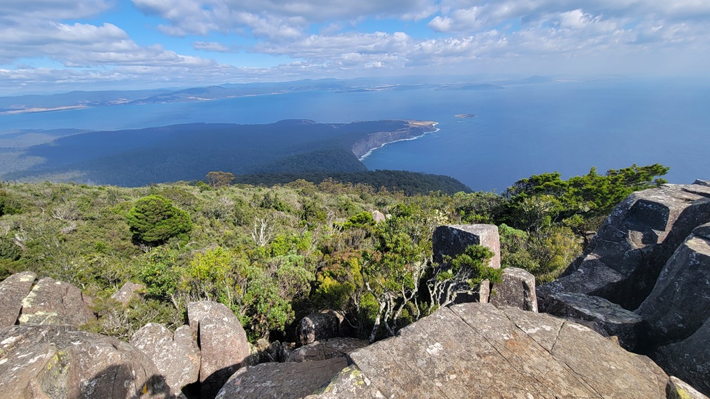 Panoramic view from the rocky summit of Bishop and Clerk on Maria Island, looking out over a rugged, forested coastline and the deep blue ocean under a bright, partly cloudy sky. In the foreground, large dolerite columns and boulders overlook a dense canopy of green native vegetation, while the distant coastline curves toward the horizon.