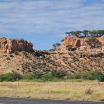 A landscape photo of two prominent red-orange sandstone mesas, known as jump-ups, rising from a grassy plain north of Winton. The flat-topped formations are crowned with green trees under a bright blue sky with scattered clouds. In the foreground, a sealed outback road and a wire fence line the edge of a golden-green field.