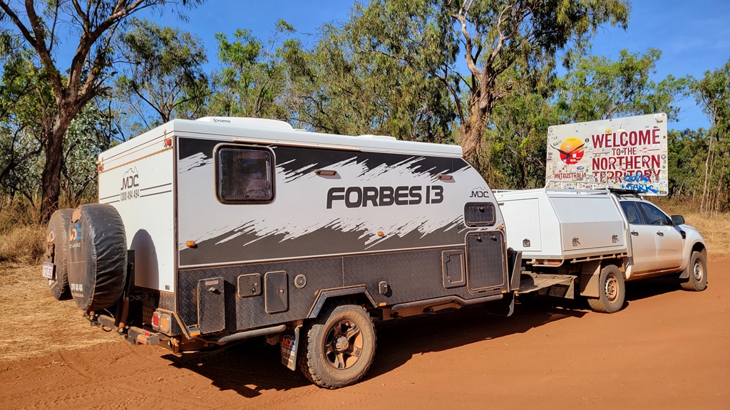 Off-grid caravanning setup featuring an MDC Forbes 13 hybrid caravan parked at the Queensland and Northern Territory border. The rig is positioned on a red dirt track typical of the Savannah Way crossing.