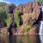 Twin waterfalls cascading down a rugged red rock escarpment into a large, dark plunge pool surrounded by lush monsoon forest at Wangi Falls, Litchfield National Park.