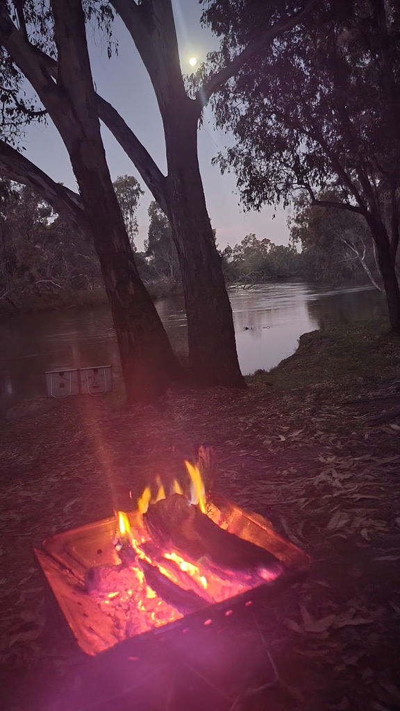 A glowing campfire burning in a portable fire pit at dusk on a Murray River road trip, with large gum trees and the quiet river visible under a twilight sky at Forges Beach.