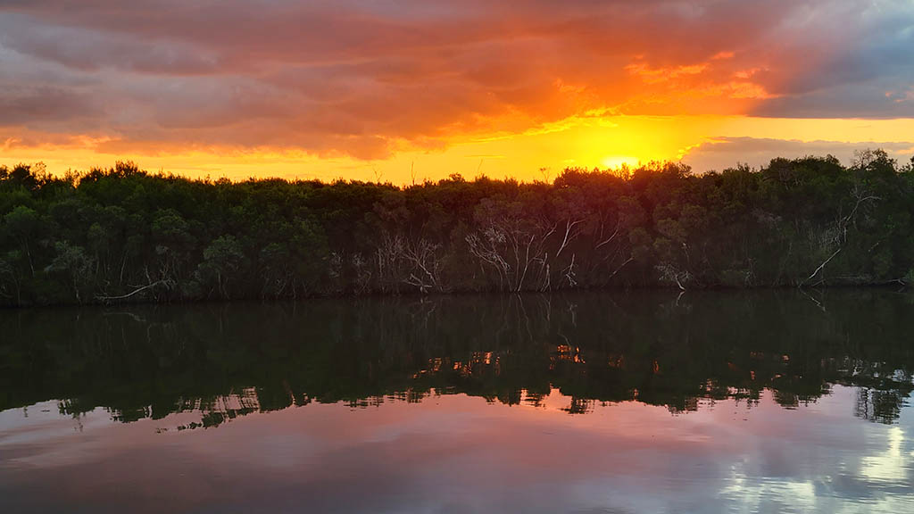 Vibrant orange sunset glowing behind treed riverbanks with calm water reflections at Fishers Riverside Campsite along the Avon River.