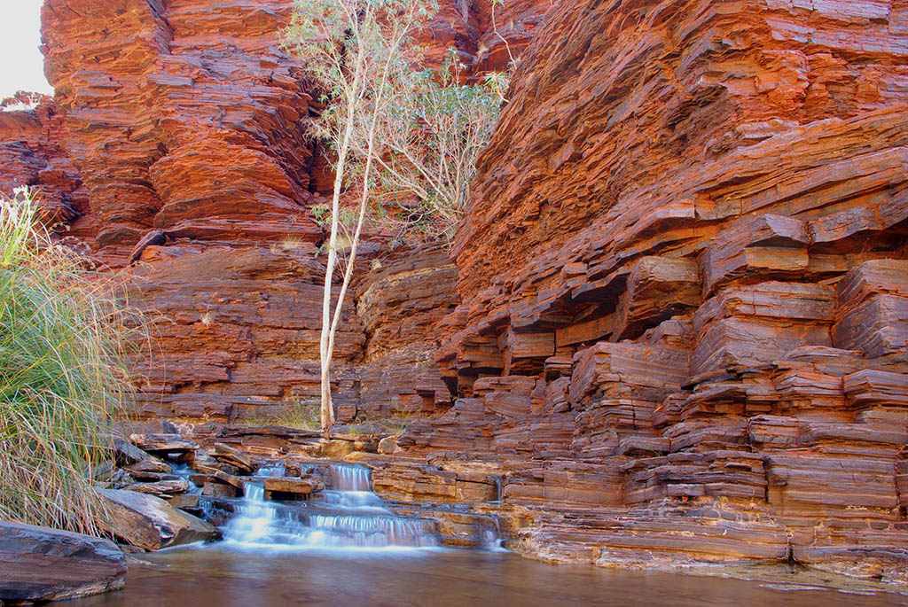 Low waterfall and rock pools framed by layered banded iron walls in Kalamina Gorge, Pilbara and Coral Coast loop